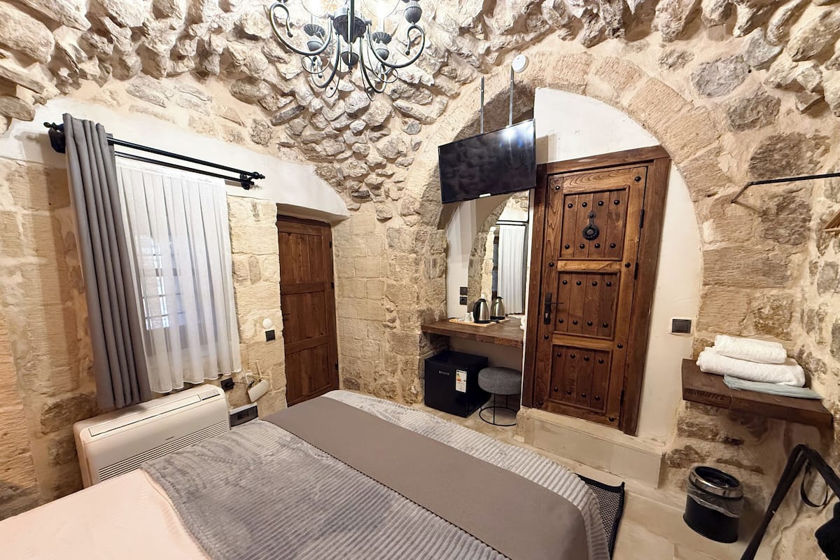 A stone-walled bedroom with a bed, a wooden wardrobe, a mirror, and a hanging TV.