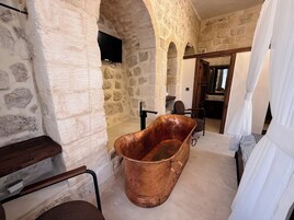 A stone-walled room with a large copper bathtub, a wooden chair, and a television mounted on the wall.