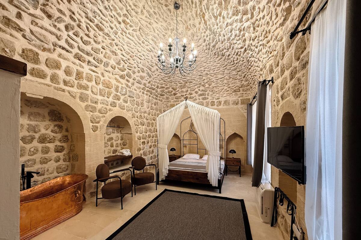 A stone-walled bedroom with a four-poster bed, a chandelier, a television, and a bathtub.