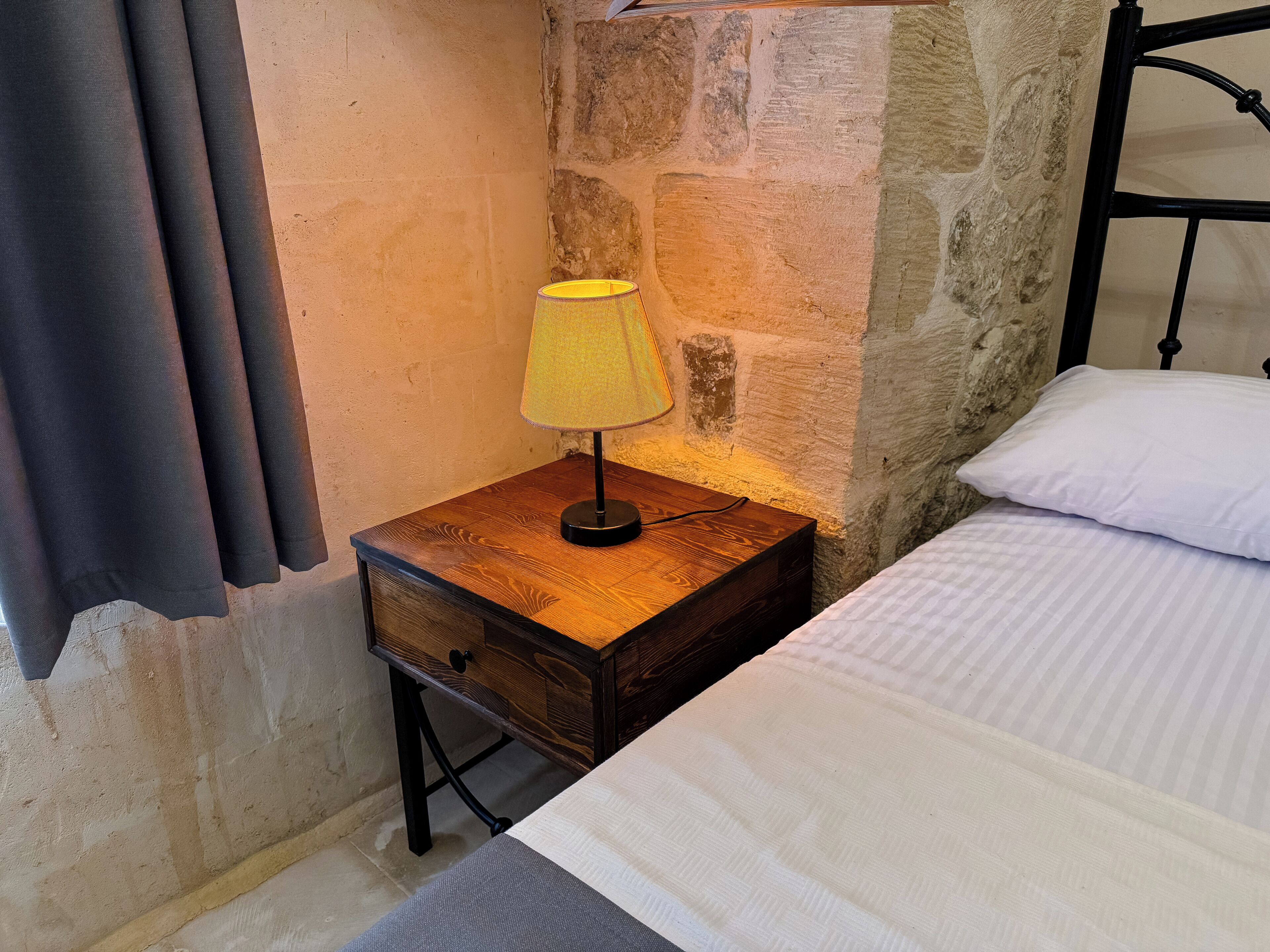 A bed with a white striped sheet, a bedside table with a lamp, and a stone wall.