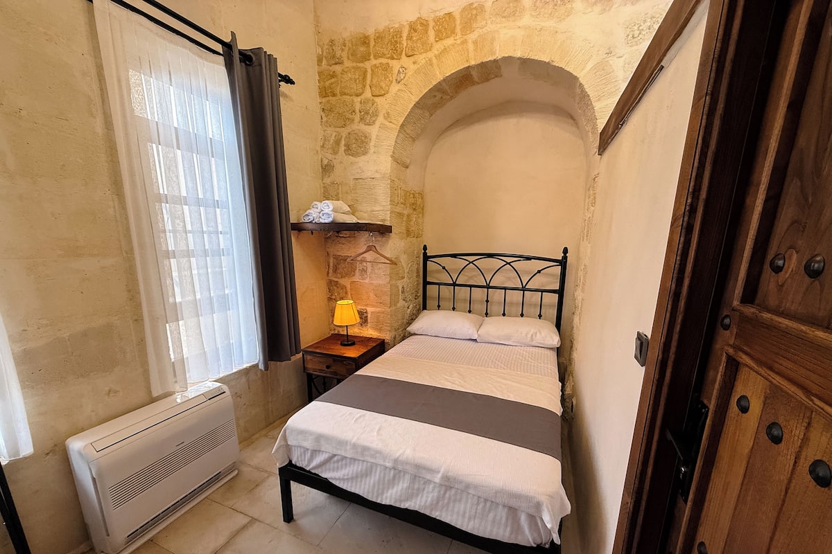 A bedroom with a stone wall, a bed with white bedding, a nightstand with a lamp, and a wooden door.