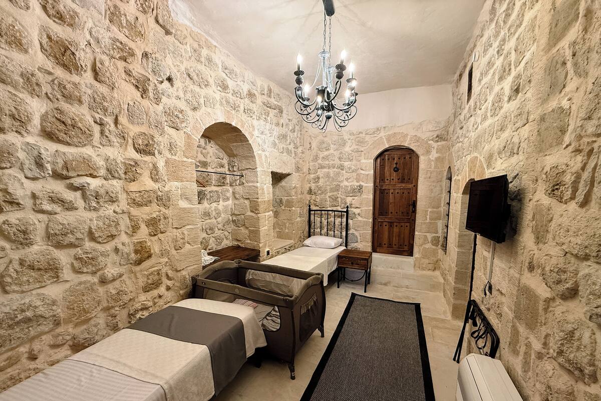 A stone-walled room with two beds, a wooden door, a chandelier, and a mounted television.
