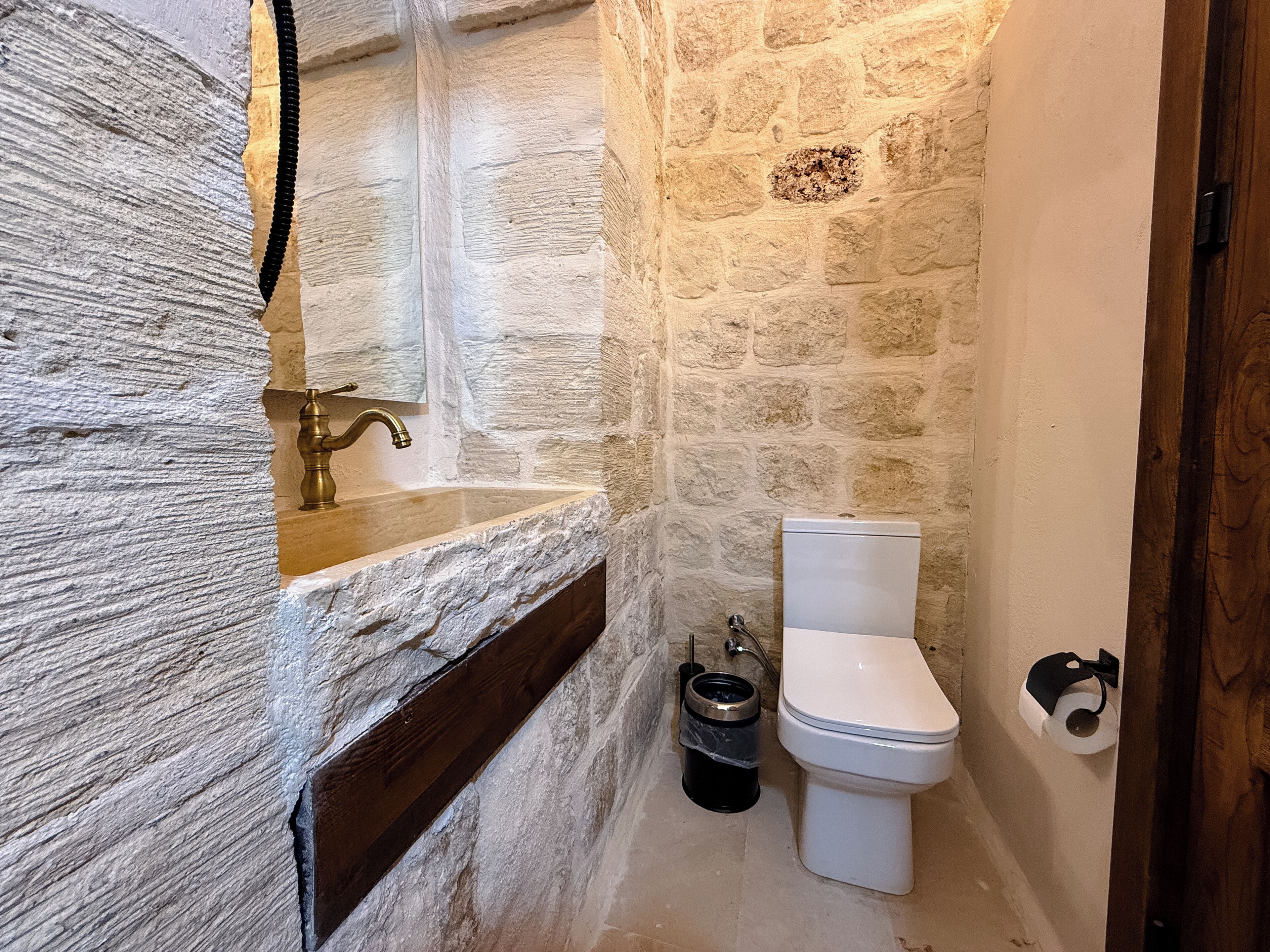 Une salle de bain avec un mur en pierre, une étagère en bois, des toilettes blanches et une poubelle noire.