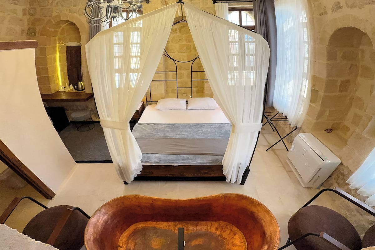 A room with a four-poster bed, a chandelier, stone walls, and a large bathtub.