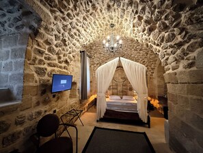 A stone-walled room with a bed under a canopy, a television on the wall, and a chair. - Sümerler Konağı (Mardin)
