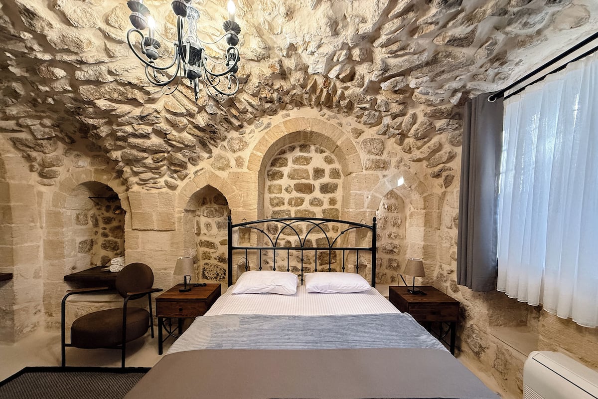 A stone-walled bedroom with a bed, two bedside tables, a chair, and a chandelier.