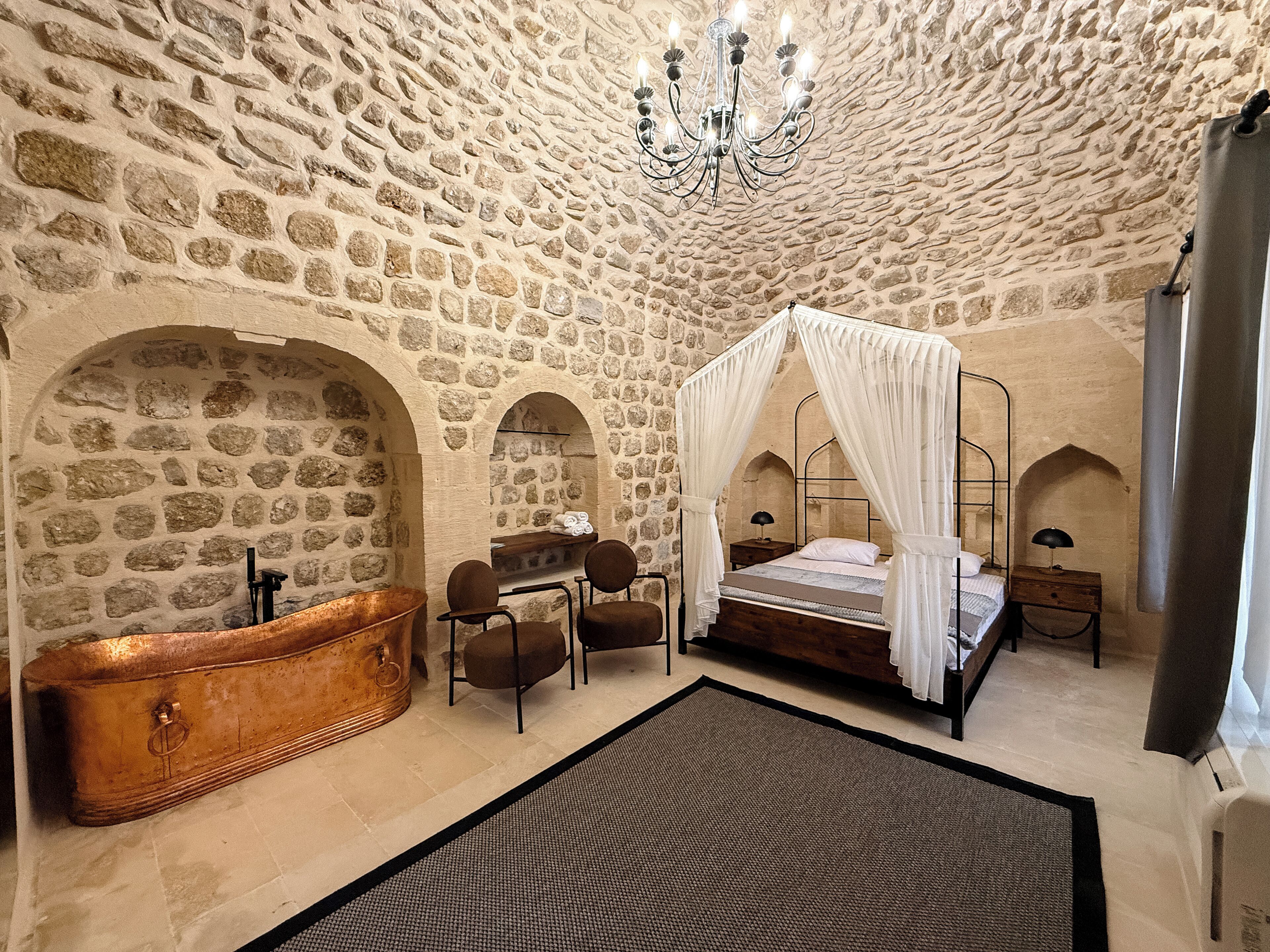 A stone-walled room with a four-poster bed, a wooden bathtub, and two armchairs.