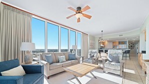 A spacious living room with a large window, a ceiling fan, a sofa, a coffee table, and a kitchen area in the background.