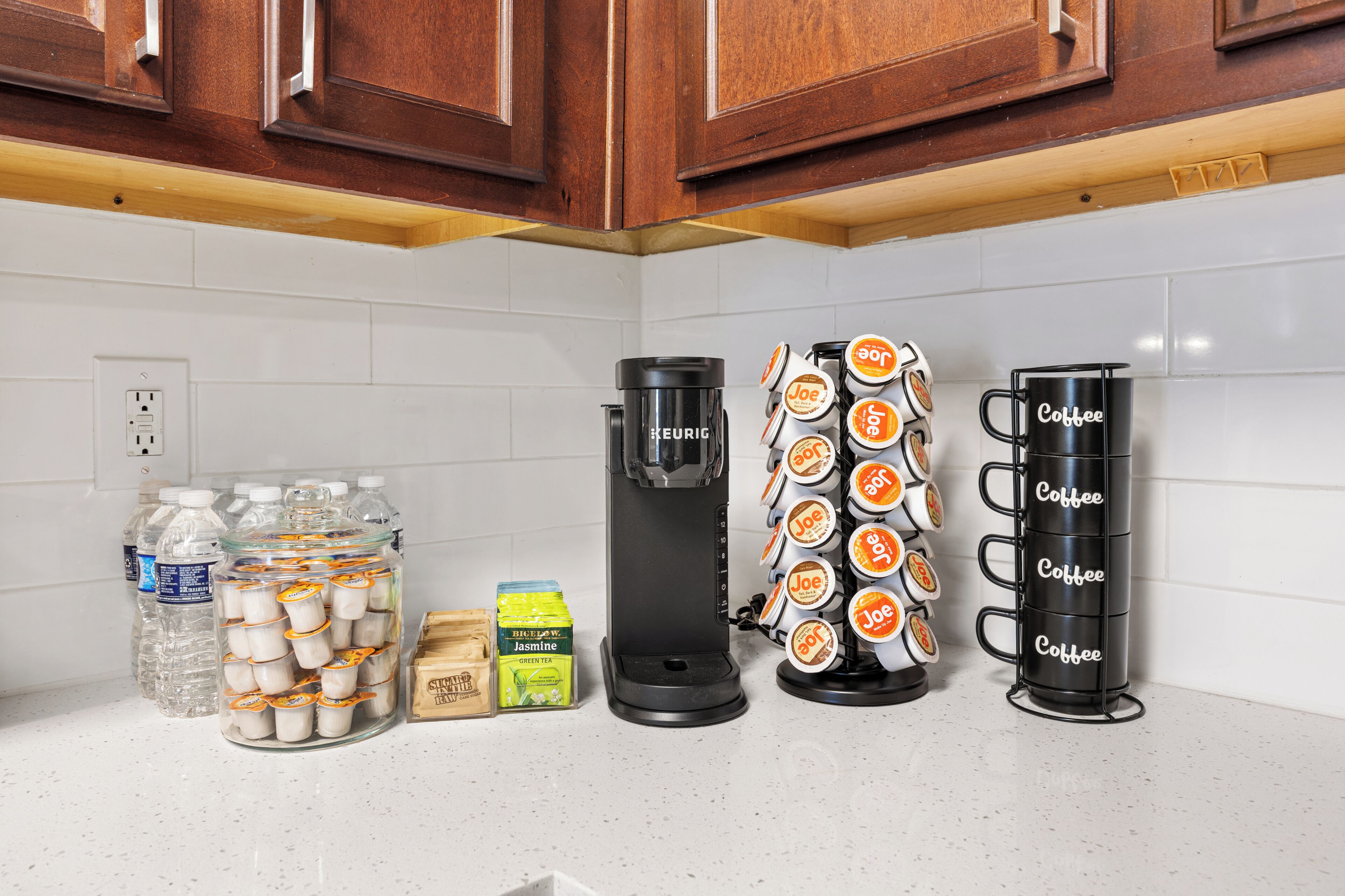 A kitchen counter with a Keurig coffee maker, a coffee mug holder, and jars of water and tea.