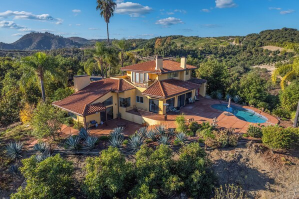 A large house with a swimming pool, surrounded by lush greenery and hills.