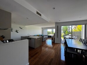 A modern kitchen with a central island, a dining area with a large table, and a view of the outdoors through large windows. - Fairway Retreat (Magenta)