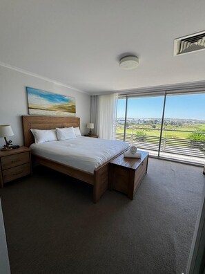 A hotel room with a large bed, a nightstand, a bedside table, a window with a view of the outdoors, and a ceiling light. - Fairway Retreat (Magenta)