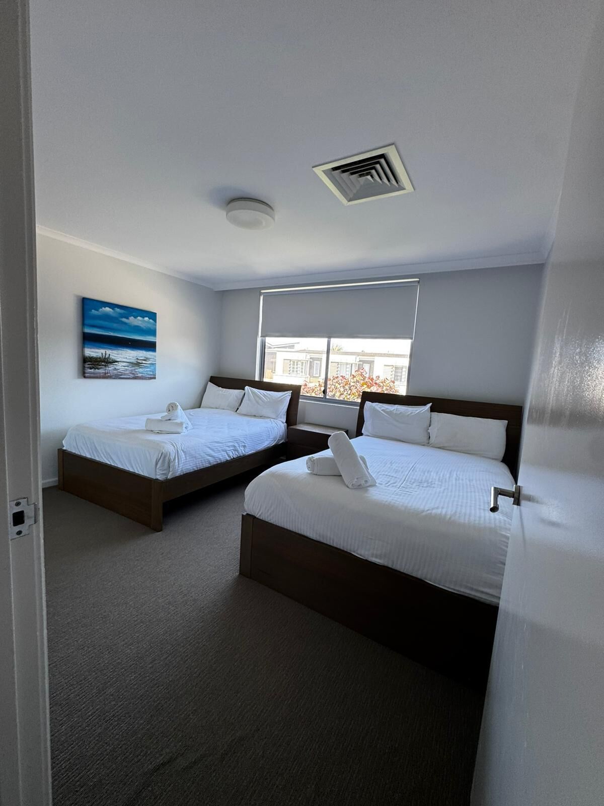 A hotel room with two beds, a painting on the wall, and a window with a view.
