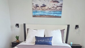Un lit bien fait, aux couleurs gris et blanc, avec un tableau représentant une scène de plage au-dessus, et un tapis à motifs noir et blanc sur le sol.