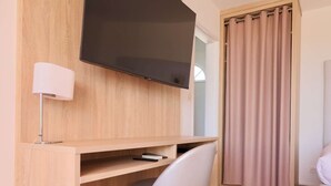 Una habitación moderna con un escritorio de madera, un televisor de pantalla plana, una silla blanca y un ventanal con cortinas.