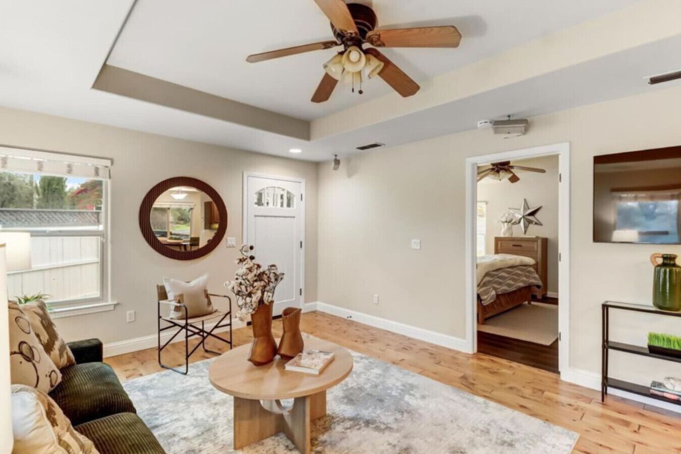 Un salon spacieux doté d’un ventilateur de plafond, d’un miroir rond, d’une table basse, d’un canapé et d’une porte donnant sur une autre pièce.