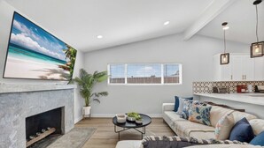 A living room with a fireplace, a large painting of a beach, a sofa, a coffee table, and a window with blinds. - Hot Tub & Canyon Views! | 10 Min to La Jolla Beach (San Diego)