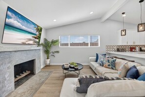 Ein Wohnzimmer mit Kamin, einem großen Strandbild, einer Couch, einem Couchtisch und einem Fenster mit Jalousien.