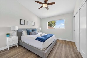 Ein Schlafzimmer mit Bett, Nachttisch, Lampe, Deckenventilator, Fenster und Blick ins Freie.