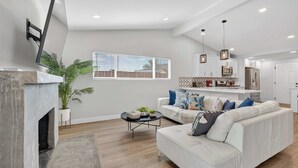 A modern living room with a fireplace, a sofa, a coffee table, and a kitchen in the background. - Hot Tub & Canyon Views! | 10 Min to La Jolla Beach (San Diego)