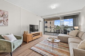 A modern living room with a sofa, a TV, a wooden side table, and a view of the city through large windows. - Luxe 3BR Apt w/ River Views + Resort Amenitie (Brisbane)