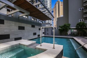 Zona de piscina con una pérgola de madera, una escalera de piscina metálica y vistas a edificios altos al fondo.
