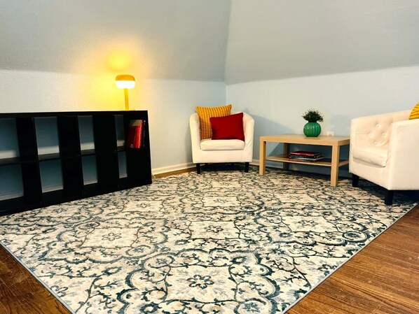 A room with a patterned carpet, a white armchair with a red pillow, a small wooden table, and a black shelving unit.