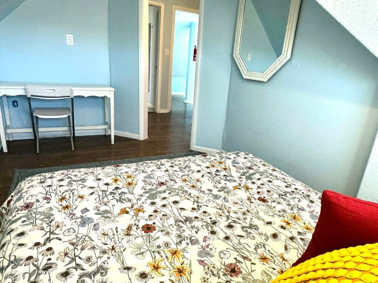 A bedroom with a floral bedspread, a red pillow, a white desk with two chairs, and a mirror on the wall.