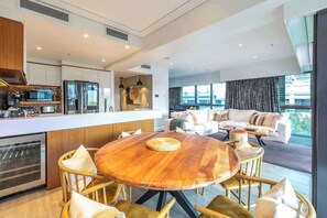 A modern kitchen with a central island, a dining area with a round wooden table, and a living room with a sofa and large windows. - Auckland CBD Viaduct Apartment - Location & Views! (Auckland CBD)