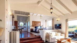 A spacious living area with a sofa, coffee table, and a television. There is a staircase leading to a bedroom with a large bed and a view of the outdoors. - The Loft at Cypress Ridge Estate (Waiheke Island)