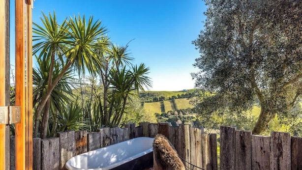 A wooden fence, a hammock, and a bathtub in a garden with palm trees and a view of rolling hills. - The Loft at Cypress Ridge Estate (Waiheke Island)