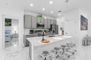 A modern kitchen with grey cabinets, stainless steel appliances, a central island with stools, and pendant lighting. - Escape to El Oasis Azul! Relaxing Cape Coral Vacation Retreat + Heated Pool. (Cape Coral)