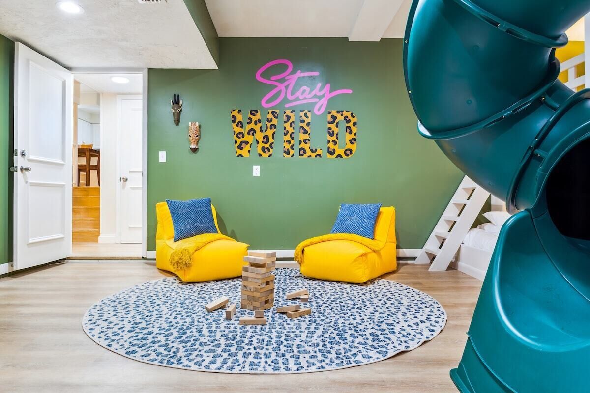 Una habitación con pared verde, dos sillas amarillas, alfombra azul y blanca estampada, y una estructura de juegos.