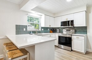 Une cuisine moderne avec des meubles blancs, un îlot central, un four à micro-ondes, une cuisinière et une fenêtre avec des stores.