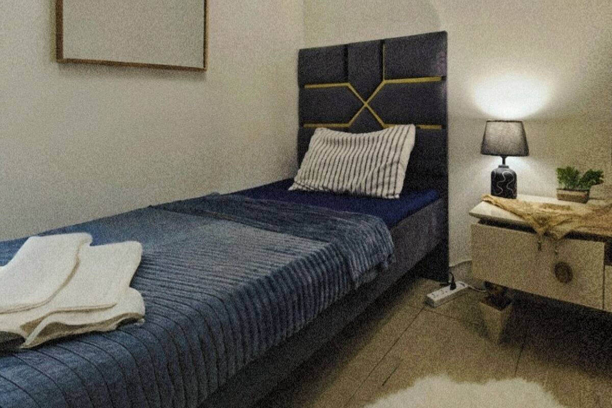 Een slaapkamer met een bed, nachtkastje, lamp en een lijst met een afbeelding aan de muur.