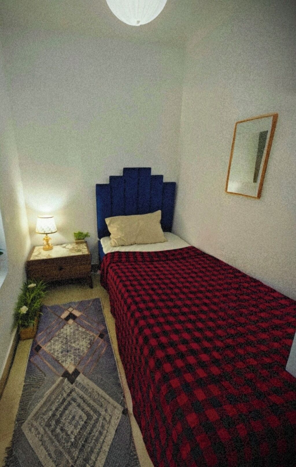 Ein kleines, aufgeräumtes Schlafzimmer mit einem Einzelbett, einem Nachttisch mit Lampe, einem Spiegel an der Wand und einem Teppich mit Muster.