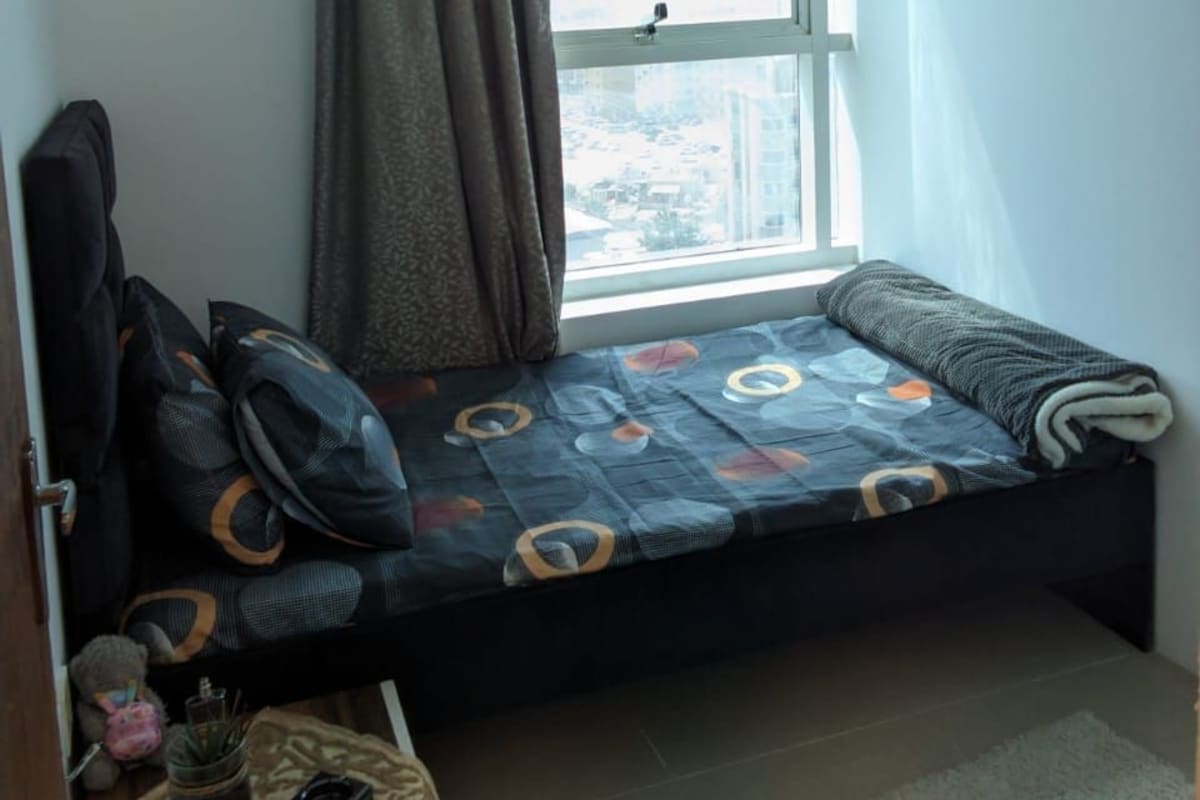 A small room with a window, a bed with patterned bedding, a small table with a bottle and a can, and a teddy bear.