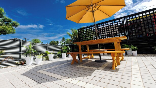 Un banc en bois sous un parasol jaune, entouré de plantes en pot et d’une terrasse carrelée.