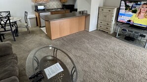 Una sala de estar con una mesa de centro de vidrio, un sofá, un televisor y un área de cocina con gabinetes de madera y un ventilador de techo.