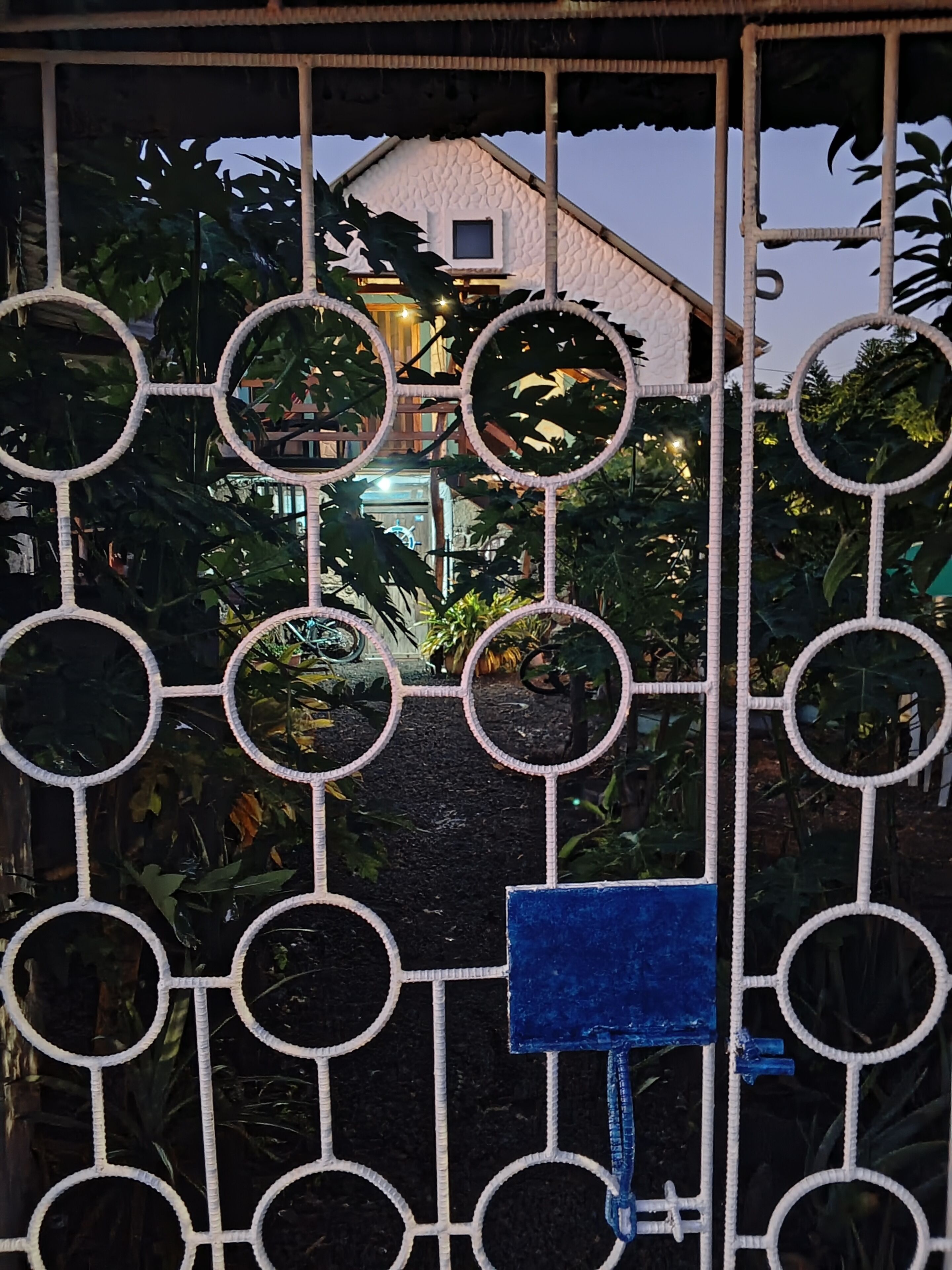 Une entrée fermée par un portail orné d’un motif géométrique en métal, un panneau bleu et, au fond, une maison dont la façade blanche présente une texture particulière.