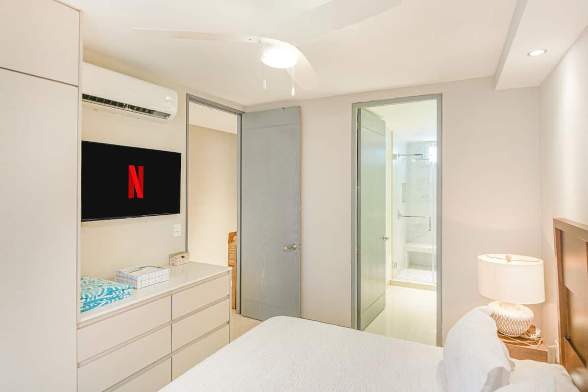 A hotel room with a bed, a TV mounted on the wall, a wardrobe, a bedside table with a lamp, and a glass door leading to a shower area.