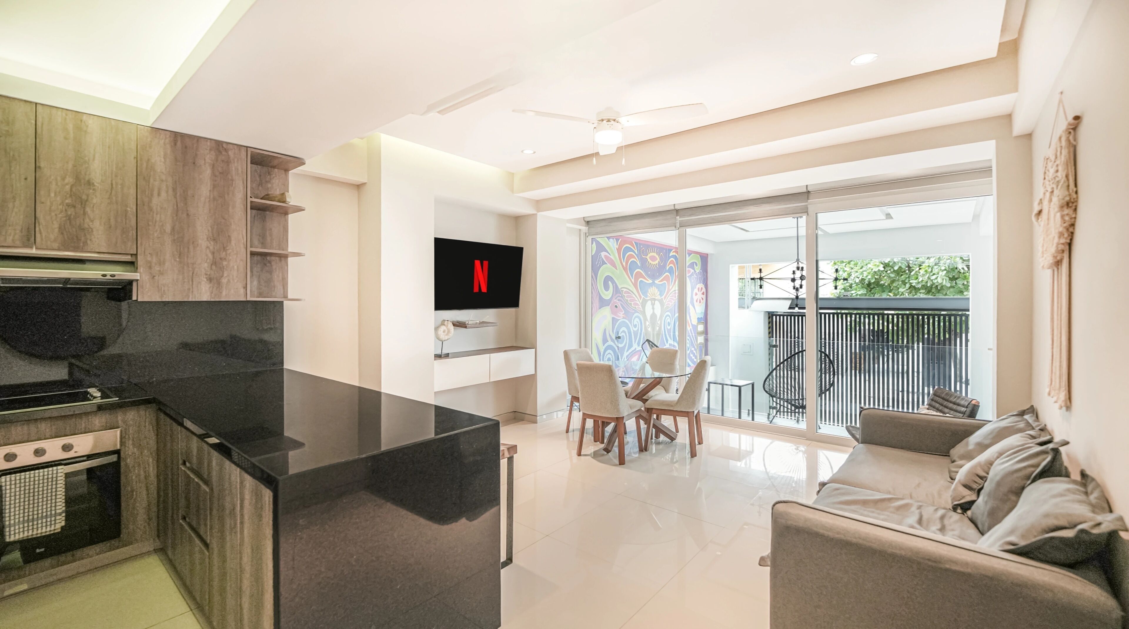 A modern kitchen with a flat-screen TV, a dining area with chairs, a sofa, and a balcony with a railing.