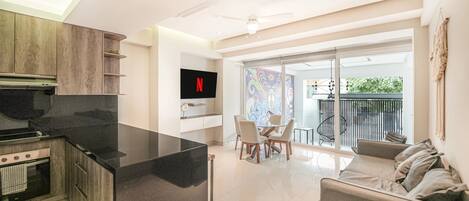 A modern kitchen with a flat-screen TV, a dining area with chairs, a sofa, and a balcony with a railing.