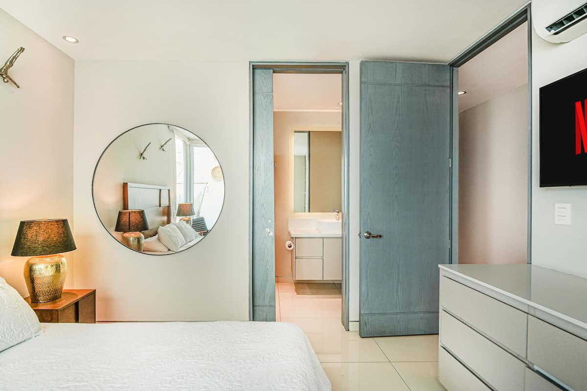 A bedroom with a bed, bedside lamp, round mirror, and a TV mounted on the wall.