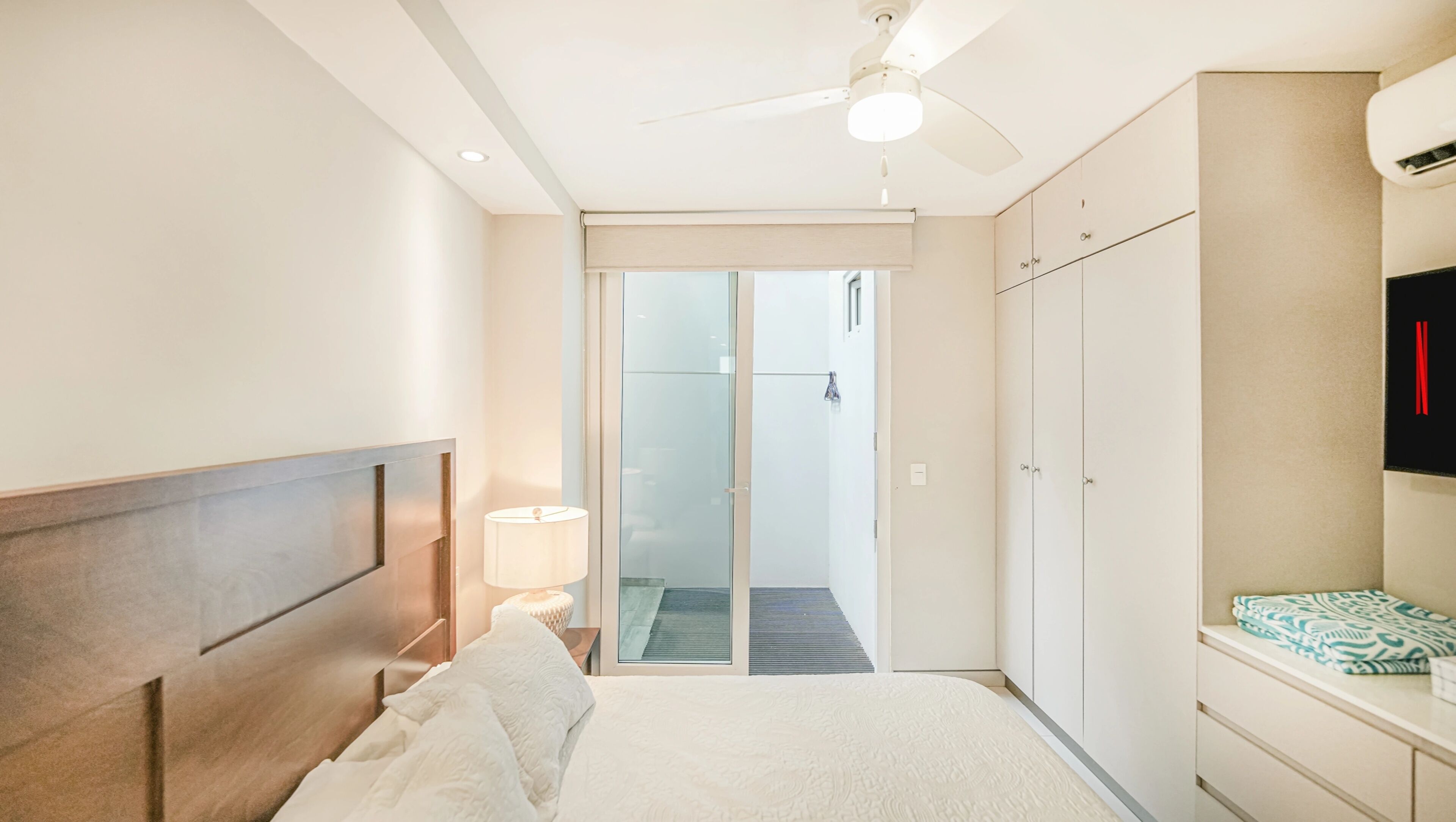 Camera d'albergo con un letto, una ventola a soffitto, una lampada, un guardaroba e una porta scorrevole in vetro che dà su un balcone.