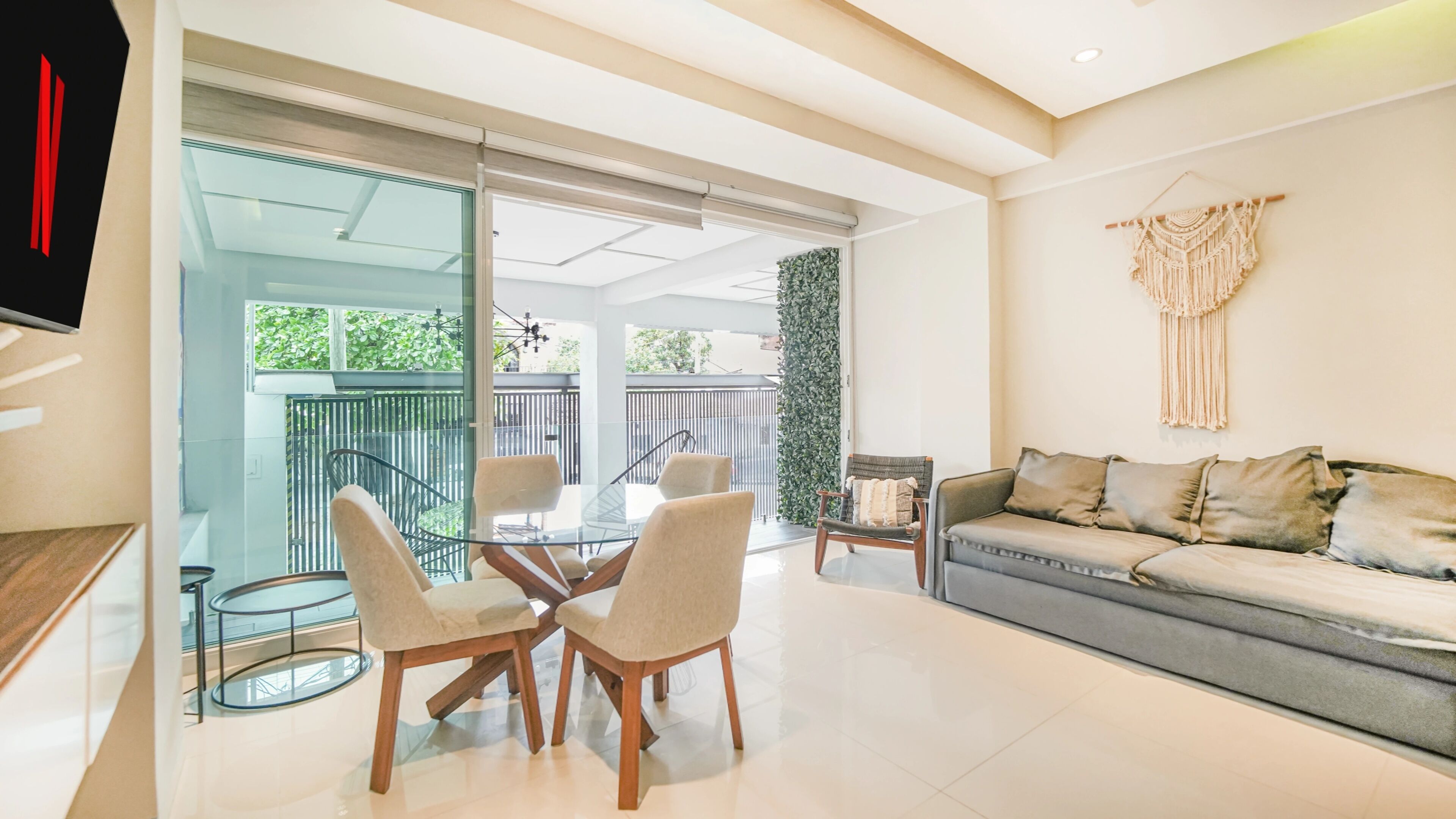 A modern living room with a glass dining table, two chairs, a sofa, and a macrame wall hanging.