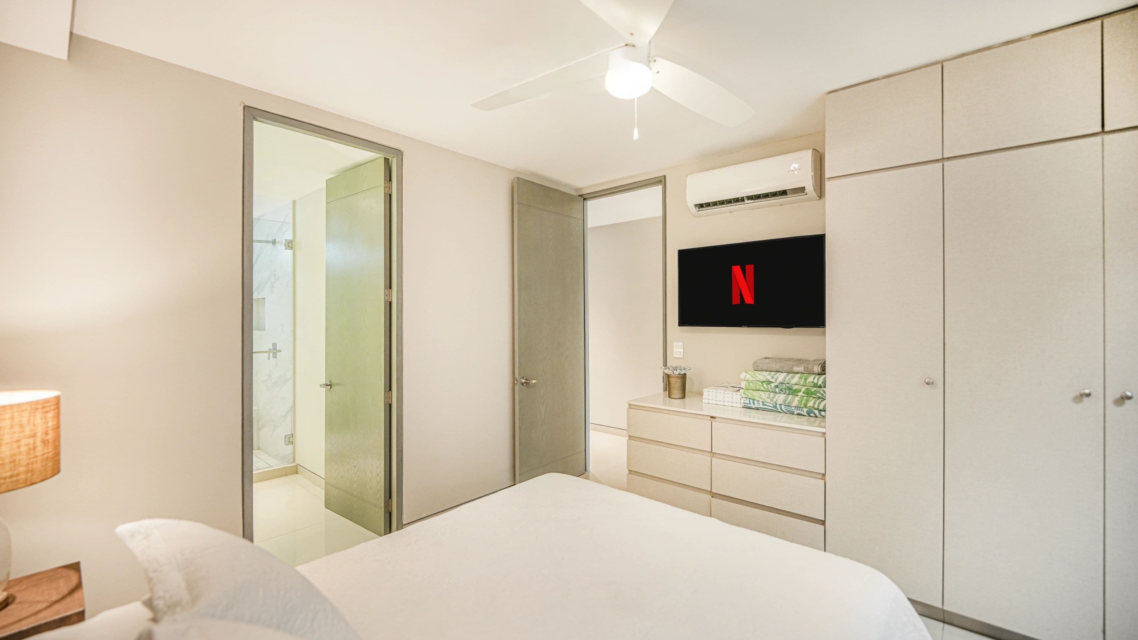 Habitación de hotel con cama, televisor montado en la pared, armario, ventilador de techo y baño visible a través de una puerta de cristal.