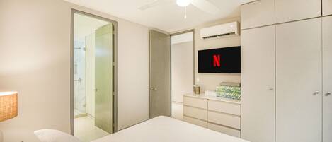 Habitación de hotel con cama, televisor montado en la pared, armario, ventilador de techo y baño visible a través de una puerta de cristal.