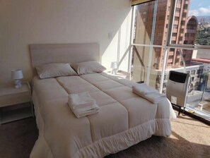 Ein großes Bett mit Kopfteil, ein Nachttisch mit Lampe und Blick auf ein Gebäude durch ein Fenster.