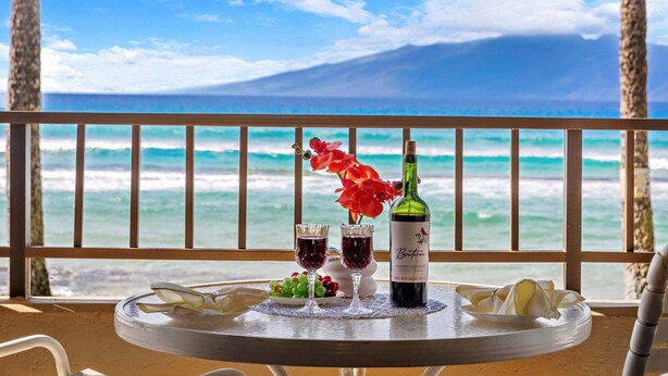 Une table avec une bouteille de vin, deux verres et un saladier de fruits, sur un balcon offrant une vue sur l’océan et une montagne.